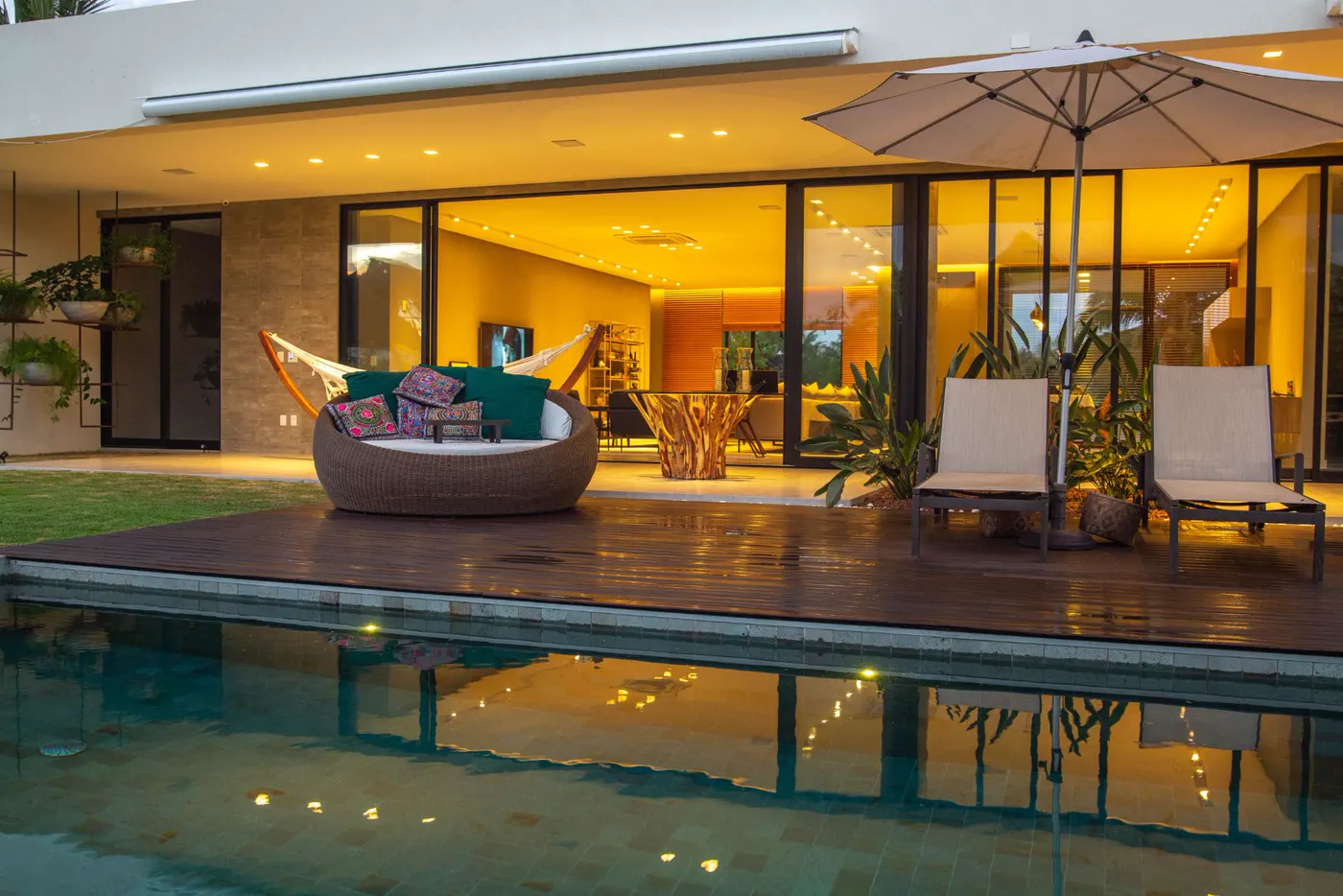Área externa ao entardecer com piscina, deck de madeira, espreguiçadeiras e a sala iluminada ao fundo